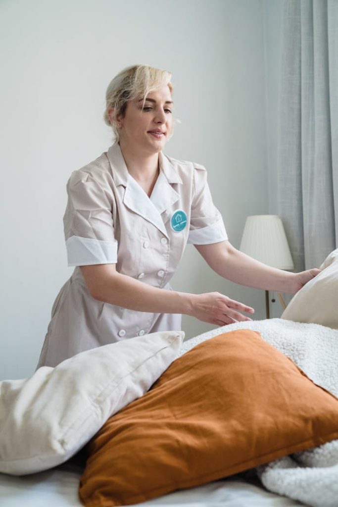 A housekeeper in uniform tidying up a bed with multiple pillows in a well-lit room.
